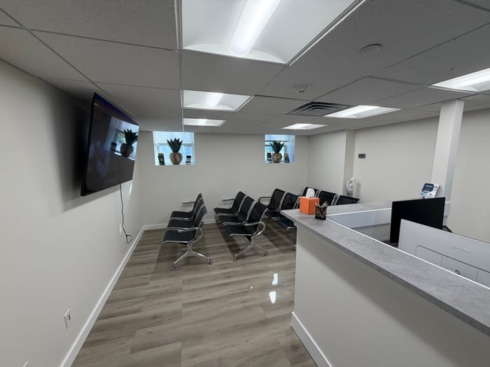 The waiting room with rows of black chairs, a wall-mounted television and two windows with plants